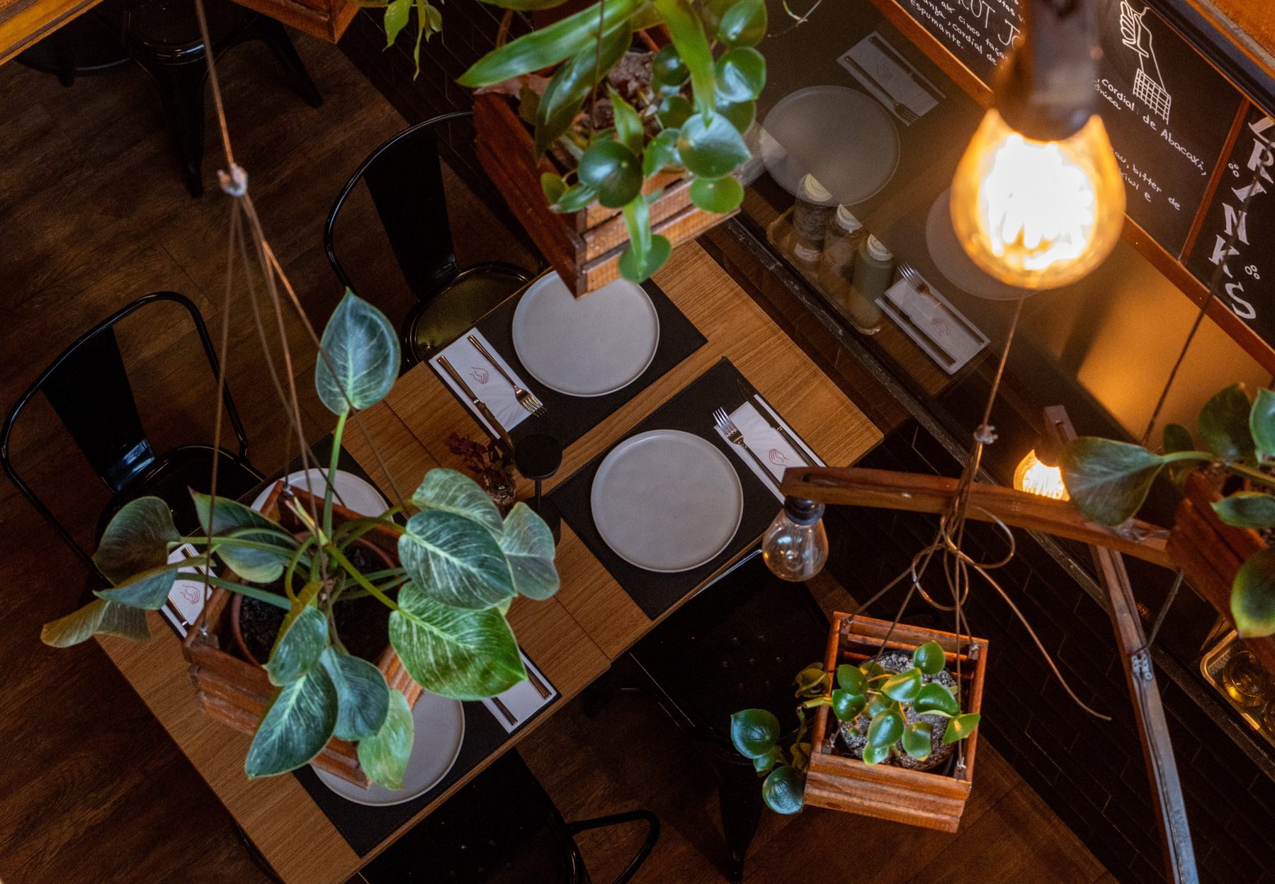 Mesas do salão com pratos, plantas penduradas em caixotes e cadeiras pretas